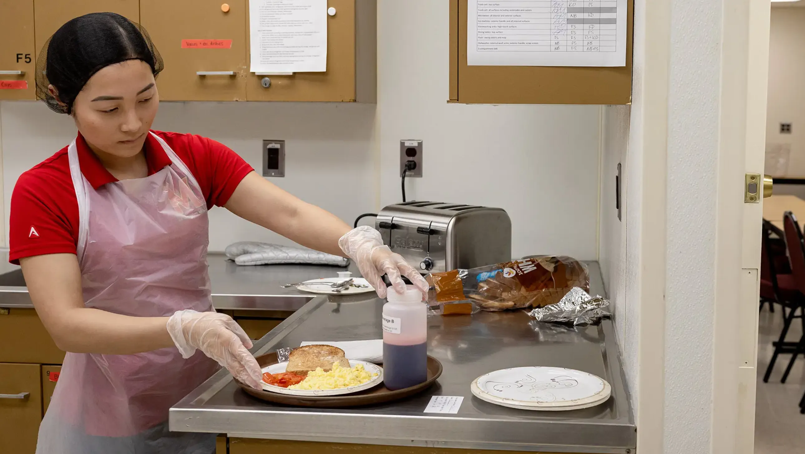 Student in a kitchen with apron and gloves, with a plate of bread, scrambled eggs and tomatoes