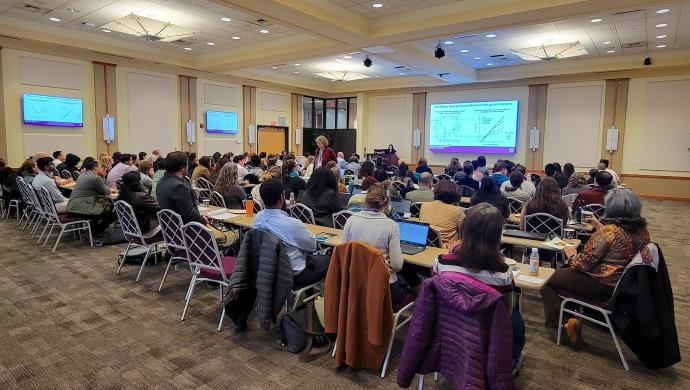 A large audience attends a microbiome research seminar, engaging with a speaker presenting on antibiotics' effects on the gut microbiome.